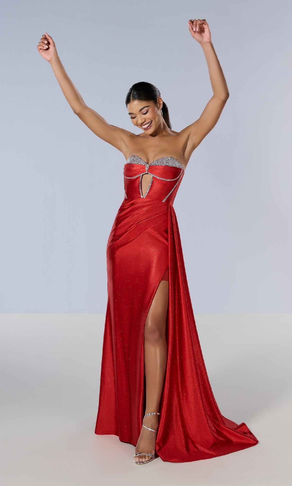 A woman in the Long Prom Dress: Tiffany 16197—a strapless glitter gown with a thigh-high slit, rhinestone accents, and a lace-up back—stands smiling with arms raised against a plain background.